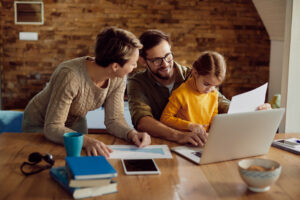 A family at a wooden table with a laptop. The child, in a yellow sweater, is focused on a paper. Parents look on, smiling and supportive. Cozy atmosphere. Title: Can You Write Off Homeschool Expenses? A Homeschooler’s 2026 Tax Guide