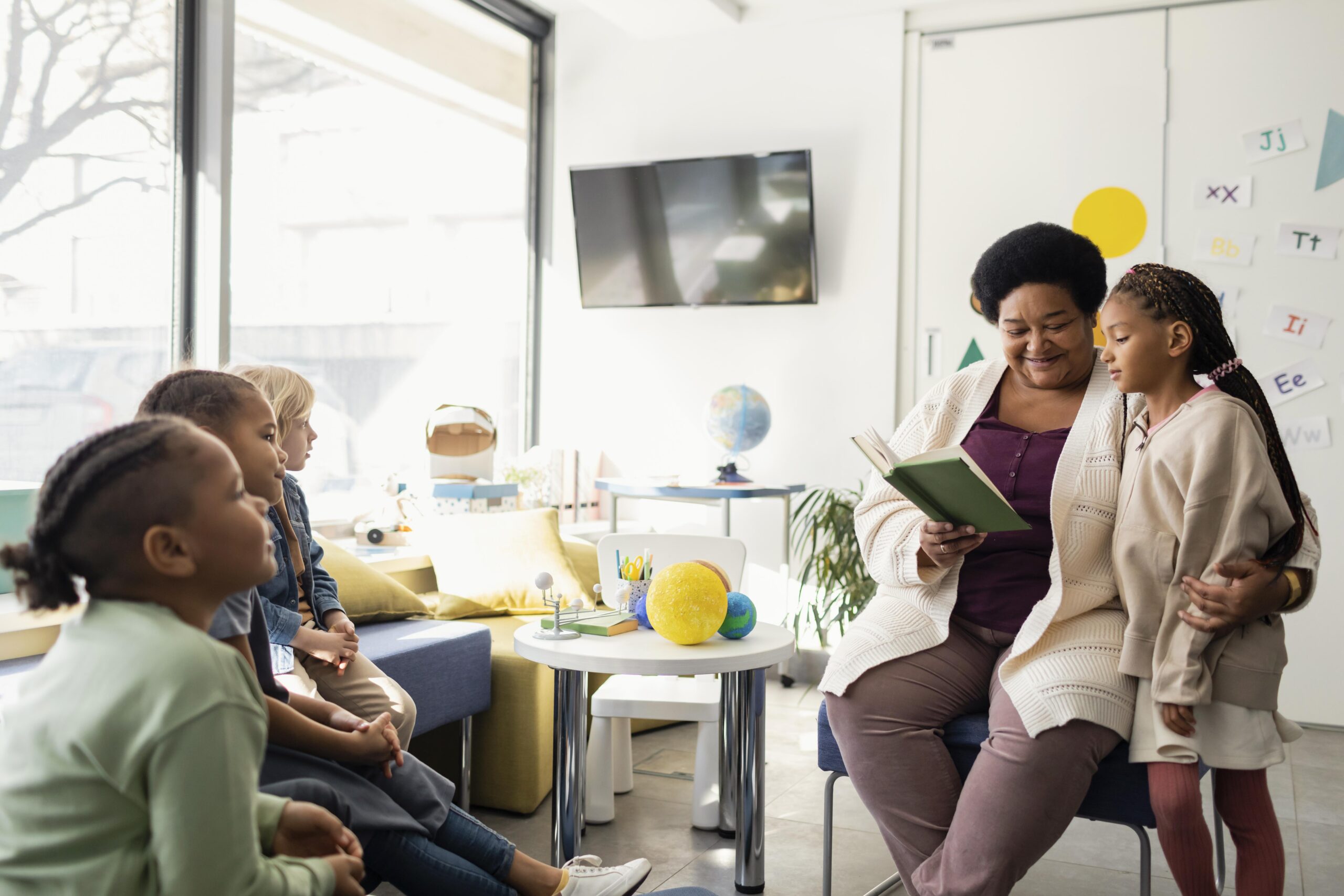 A microschool teacher reads to attentive children in a bright homeschool classroom. A girl sits close, showing engagement. The room has educational decor and natural light.