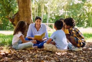 homeschool teacher reading outside under tree with children. A group of children sits with their teacher outside, reading and exploring nature together. This scene highlights outdoor learning homeschool ideas, showing how Earth Month homeschool activities can combine reading, hands-on exploration, and connection to the natural world. Perfect for inspiring backyard nature learning activities and homeschool nature study ideas.
