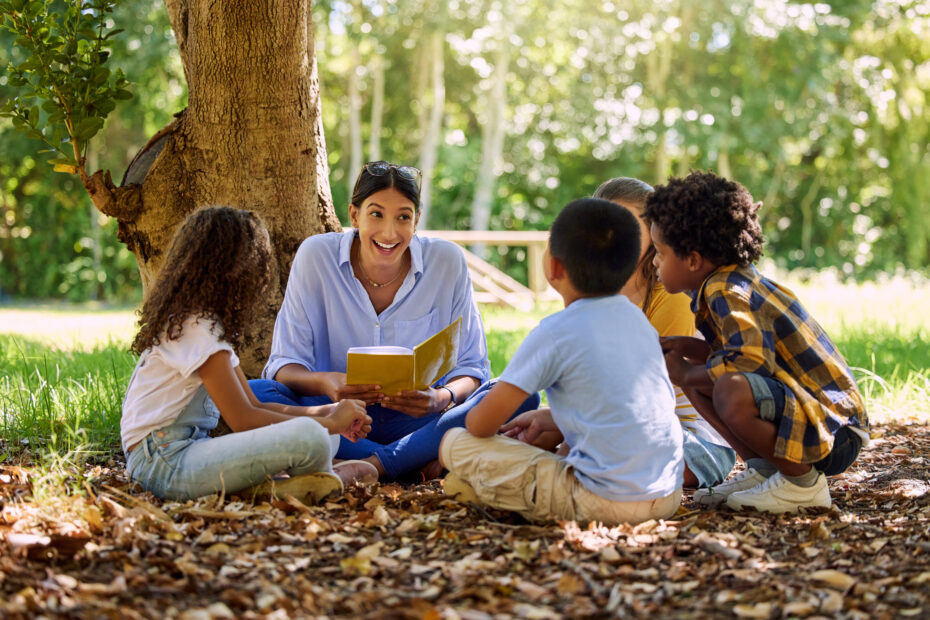 homeschool teacher reading outside under tree with children. A group of children sits with their teacher outside, reading and exploring nature together. This scene highlights outdoor learning homeschool ideas, showing how Earth Month homeschool activities can combine reading, hands-on exploration, and connection to the natural world. Perfect for inspiring backyard nature learning activities and homeschool nature study ideas.