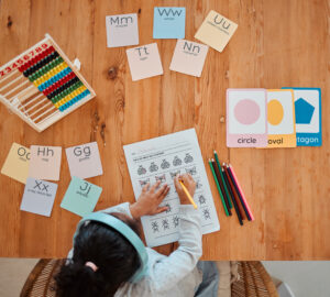 Child working on a math worksheet with colorful pencils, letter and shape cards, and an abacus on a wooden table. Text overlay: "9 Ways to Grow Your Homeschoolers Love of Math."
