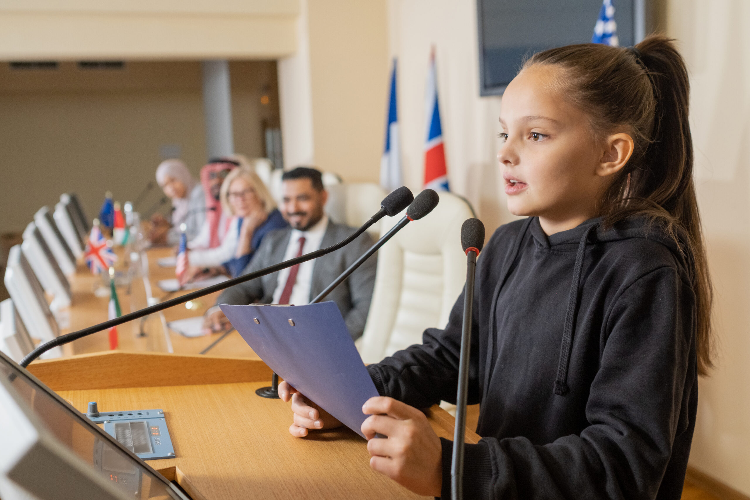 A young girl in a black hoodie speaks confidently at a podium with microphones. Diverse international delegates sit behind her, with multiple flags visible. What Connecticut's House Bill 5468 Could Mean for Homeschooling Nationwide