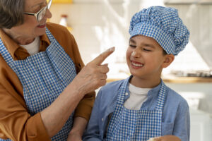 Homeschooled child in a blue baker uniform smiling while a parent playfully dabs whipped cream on his nose during a connected learning homeschool baking activity that encourages hands-on, interest-led education and real-world learning.
