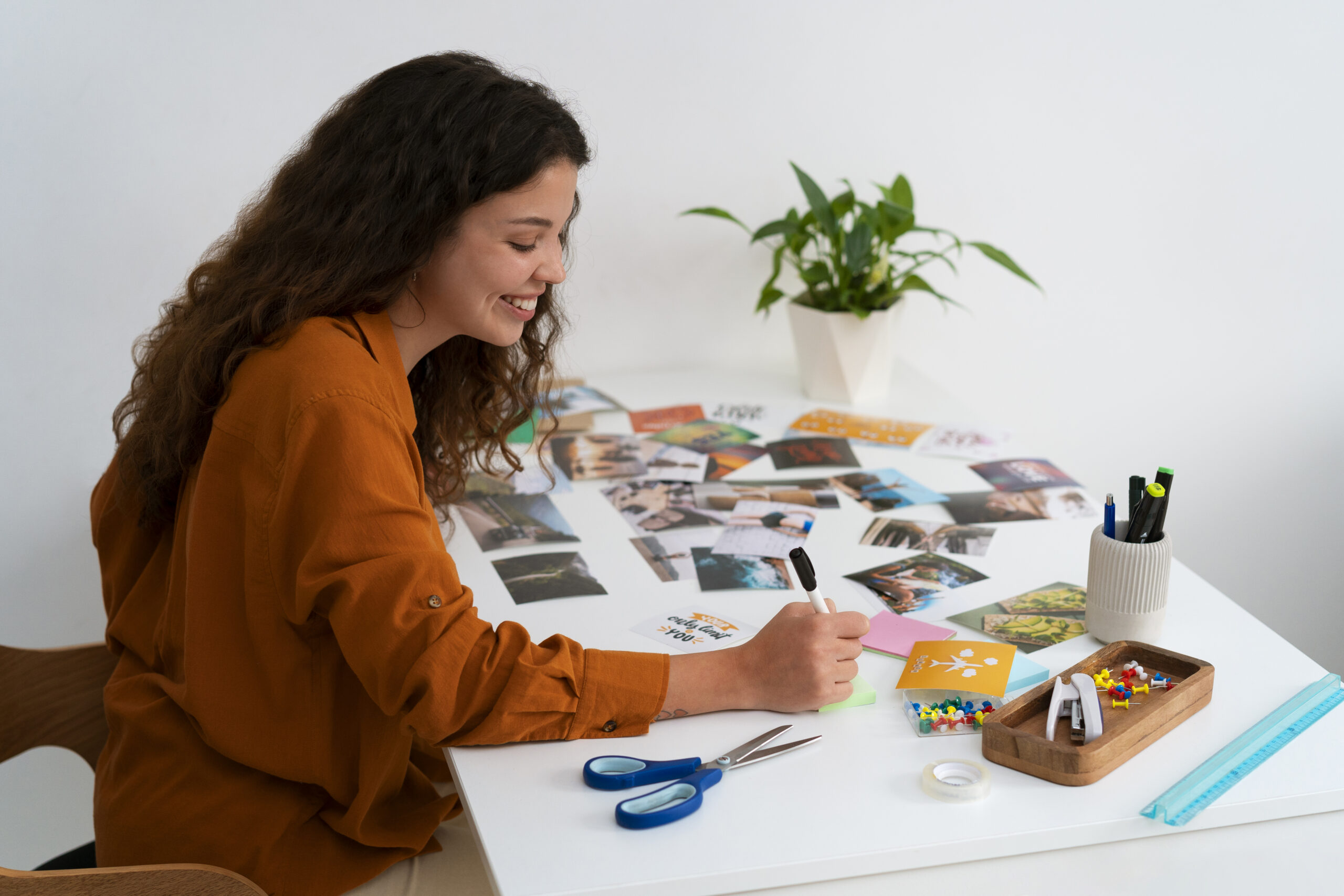 A smiling woman in an orange shirt sits at a table, organizing colorful photos, scissors, and stationery. A plant is in the background. The scene is creative and inspiring. A pink banner reads, "How to Start a Microschool: Finding Your Niche and Defining Your Vision." the women is writing steps to start a microschool