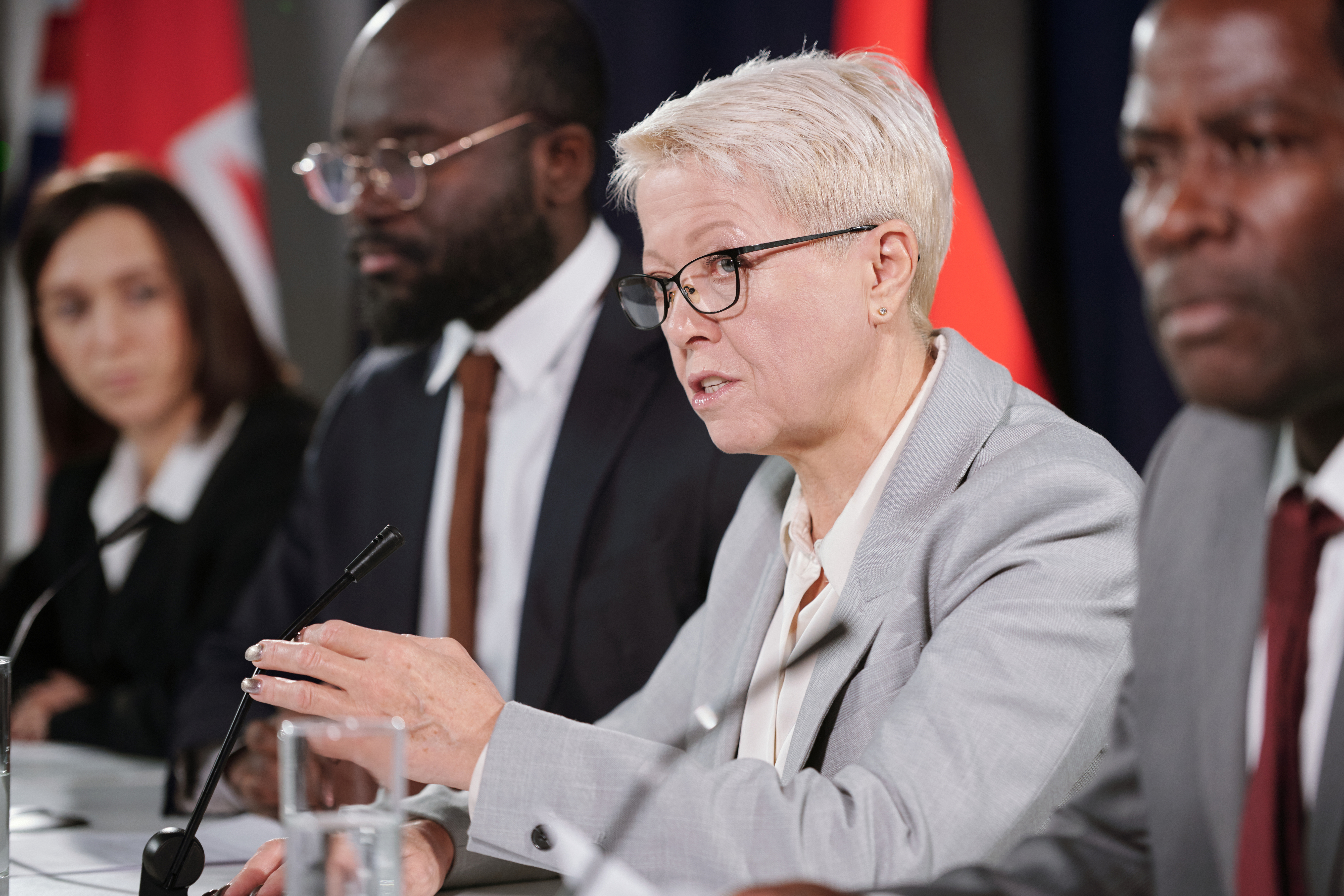 Woman talks into microphone at a panel of speakers. She is grey haired and wearing a grey suit. She looks decisive.